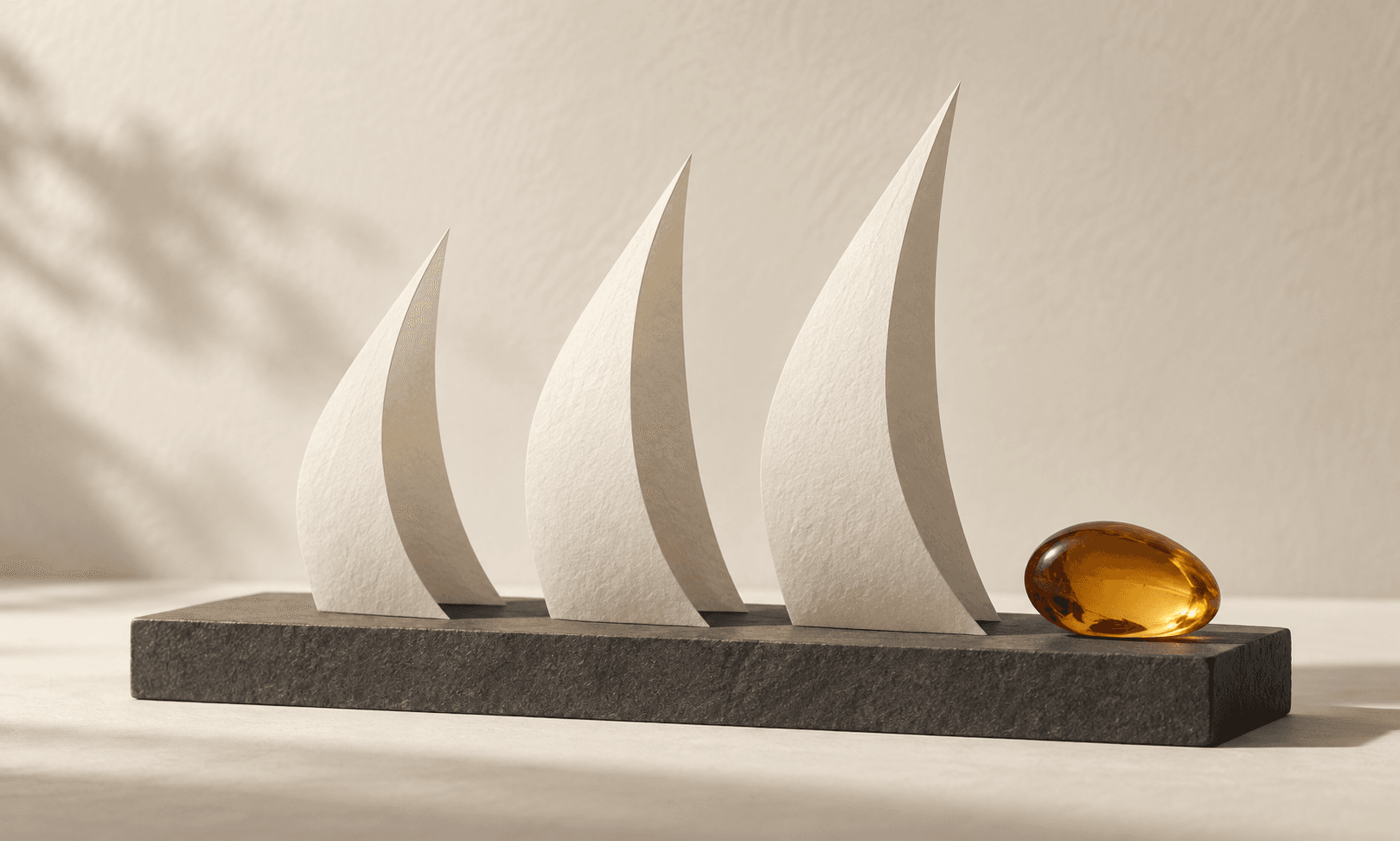 Three ivory paper sails stand on a charcoal base beside an amber pebble