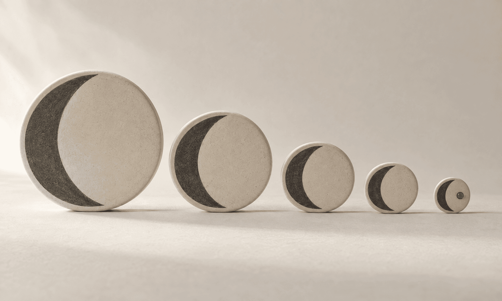A row of ivory ceramic discs shrinking from large to tiny, each inlaid with the same charcoal crescent, the smallest marked with an amber dot
