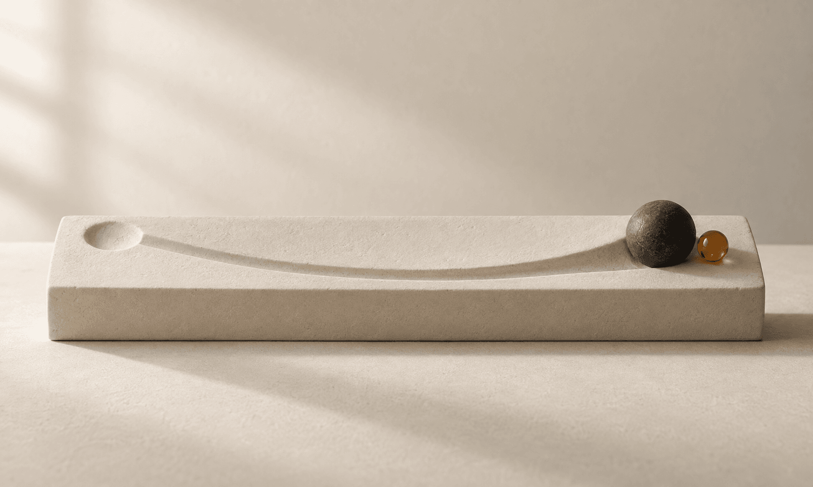 A charcoal sphere at the end of a curved groove in an ivory ceramic plinth, with a faint impression where it started and an amber bead beside it