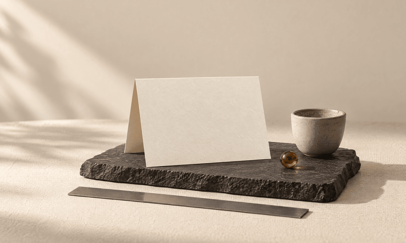 A folded blank ivory card standing on a charcoal slate beside a small ceramic cup, with a graphite ruler in front