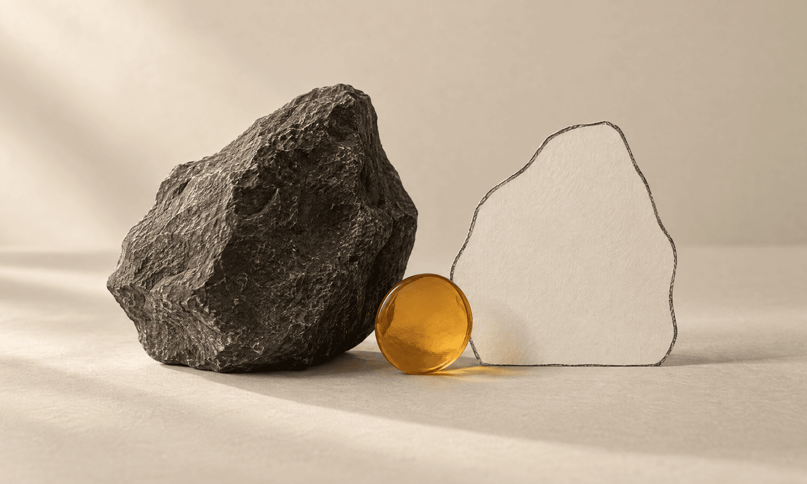 A rough charcoal stone beside an ivory paper cut-out of the same shape with a thin graphite outline, and an amber disc between them