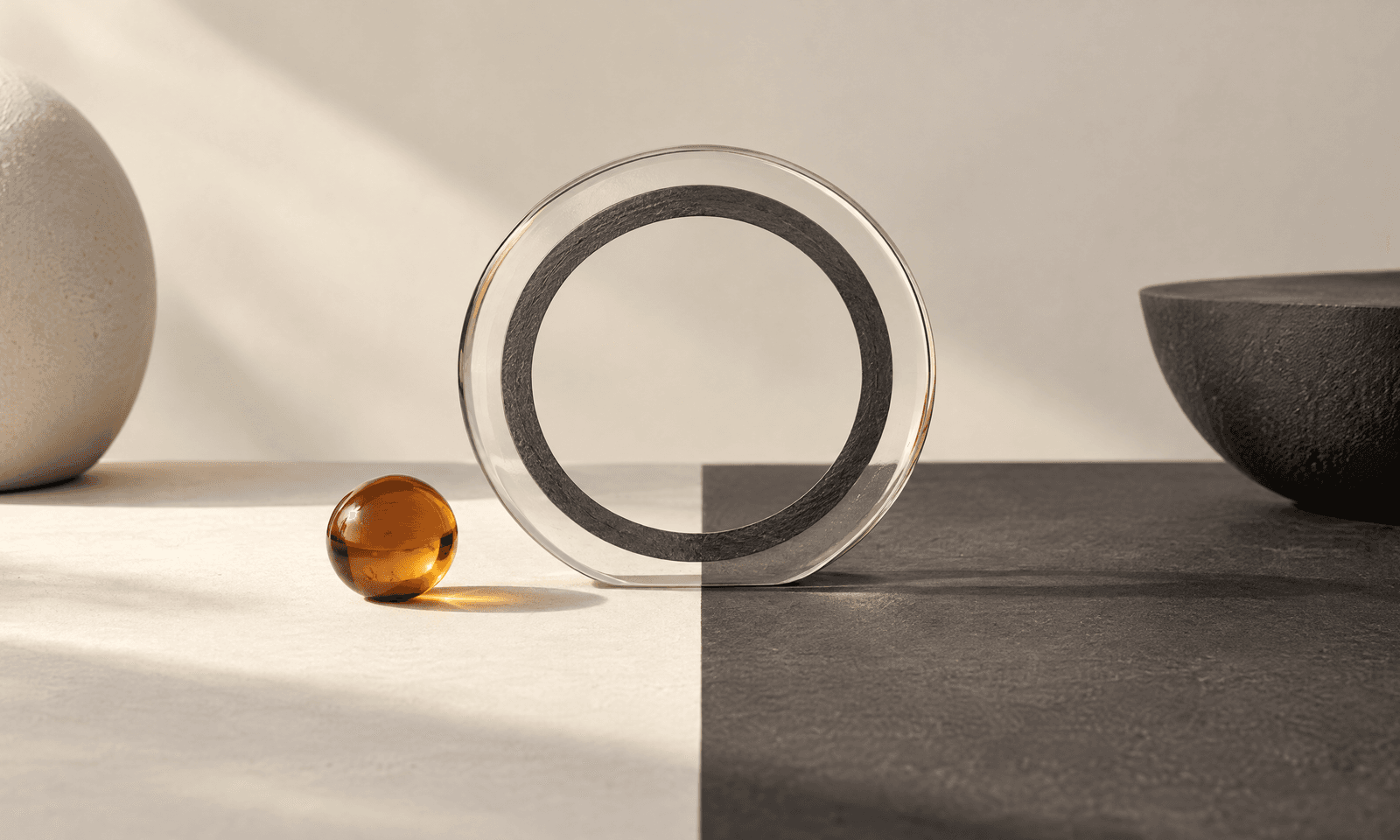 A clear glass disc with a thin charcoal ring standing on a surface split into ivory and charcoal halves, with an amber sphere nearby