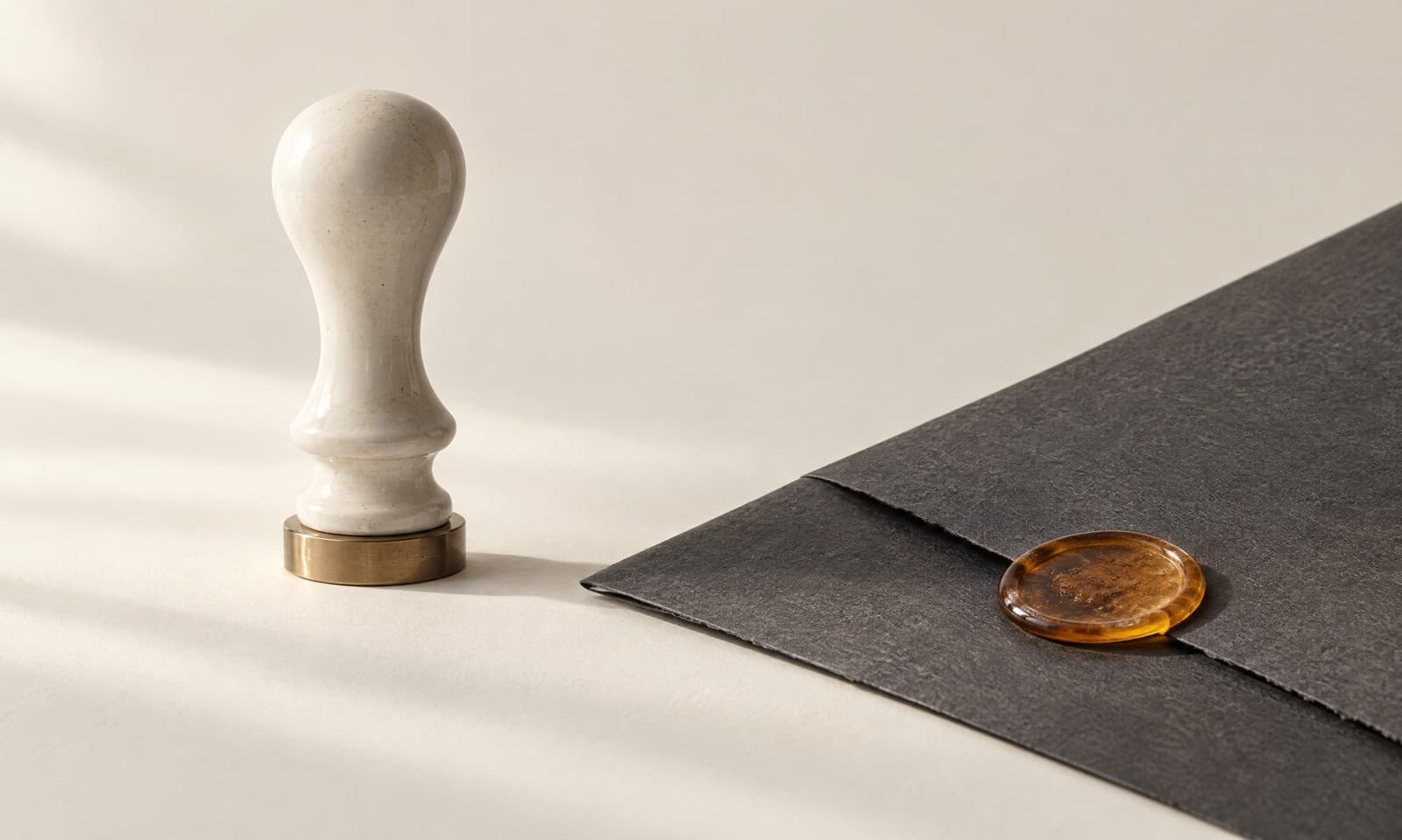 An ivory ceramic seal press standing beside a folded charcoal paper sheet with an amber wax disc pressed into its corner