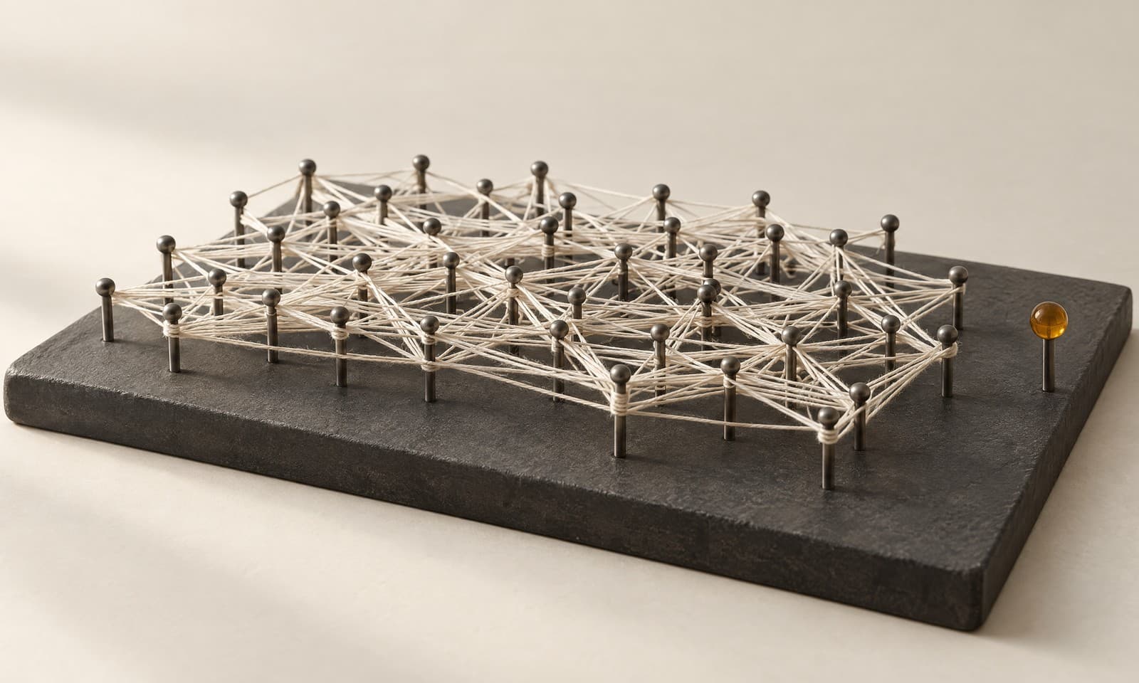 A charcoal board studded with fine graphite pins wrapped in a dense ivory thread, with one amber pin standing clear