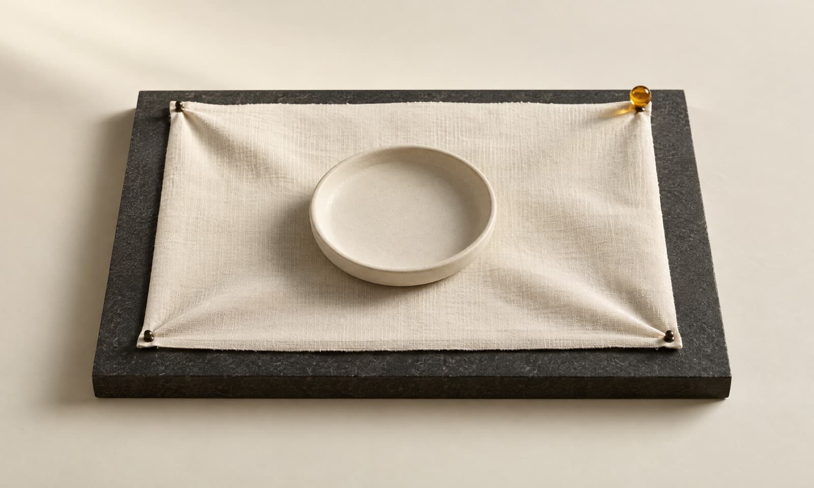 A panel of plain ivory fabric pinned flat across a charcoal board with a ceramic disc at its centre and an amber pin at the corner