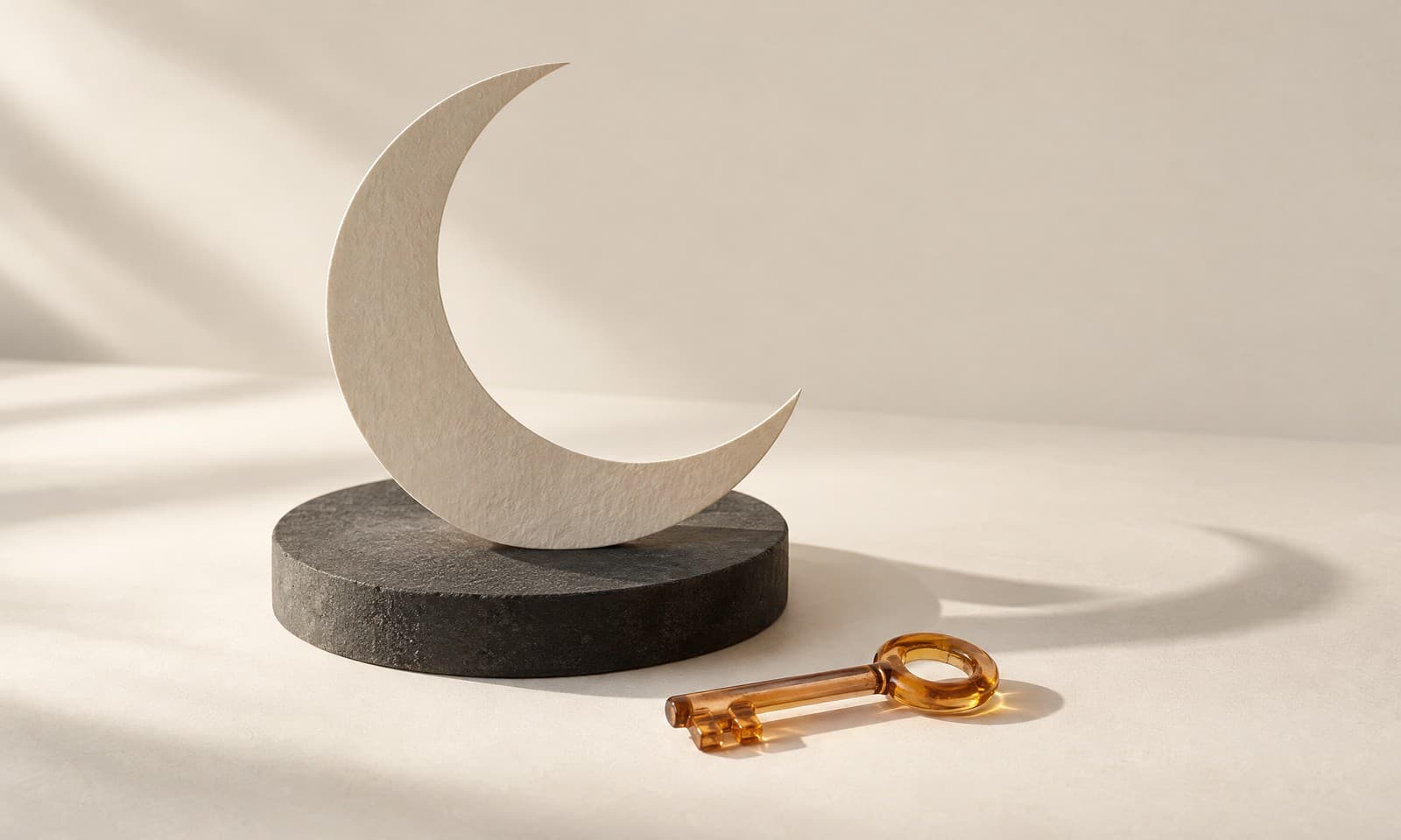 A thin ivory paper crescent standing on a charcoal disc with a small amber glass key lying in front of it