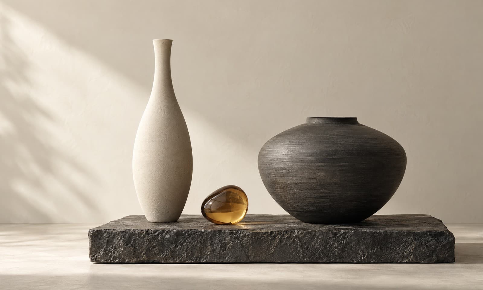 Two sculptural vessels on a charcoal slab, one tall and slender in ivory, one wide in graphite, with a drop of amber glass between them