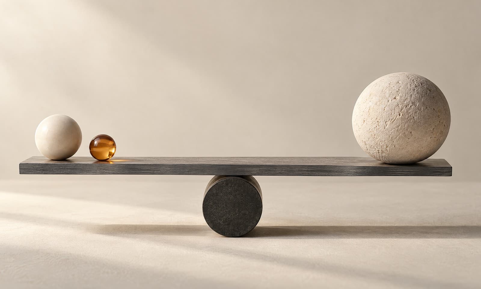 Una barra de equilibrio de grafito que sostiene una pequeña esfera de cerámica marfil y una esfera de piedra texturizada más grande, con una cuenta de vidrio ámbar junto a la pequeña