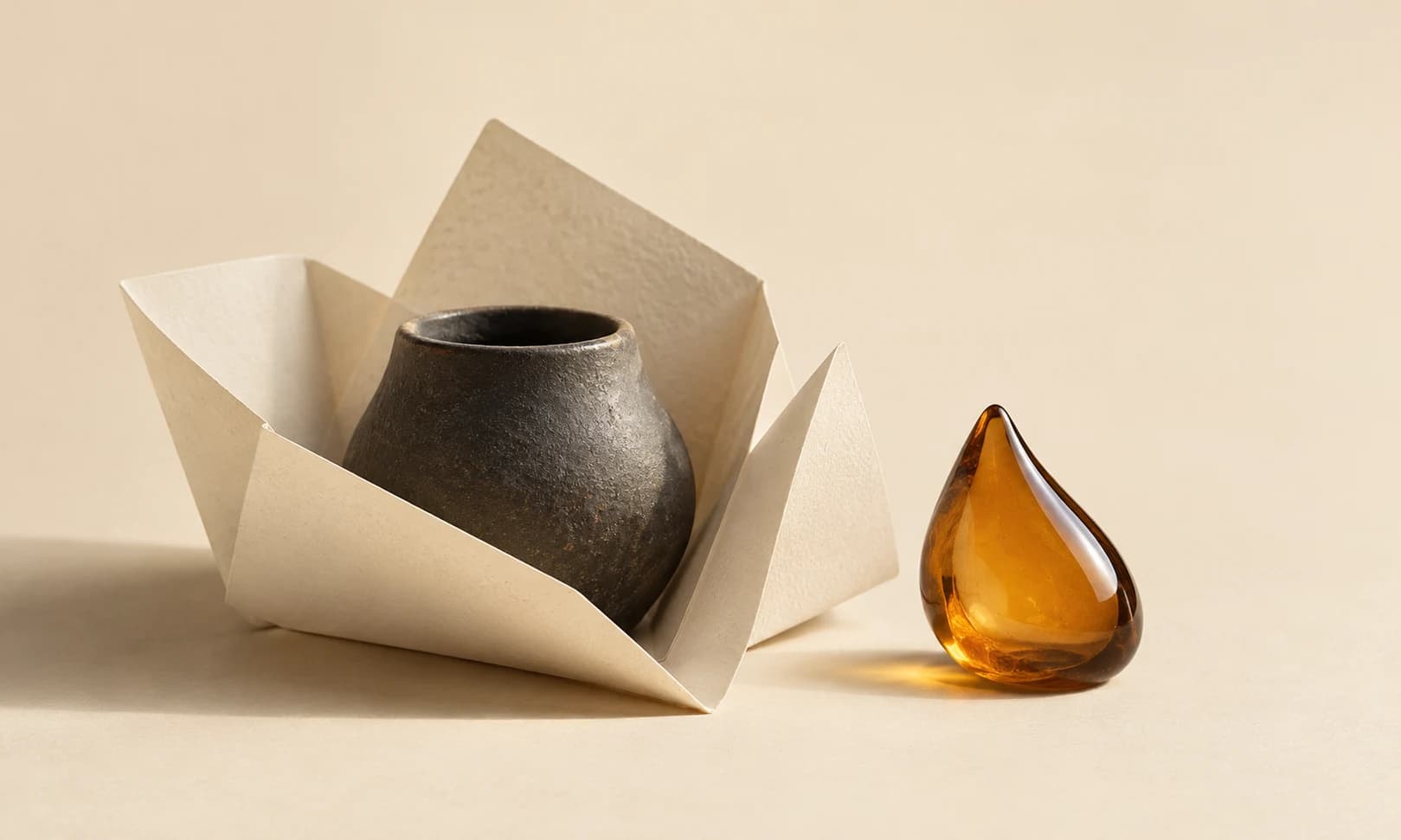 Charcoal vessel nestled in an ivory folded leaflet beside an amber droplet sculpture