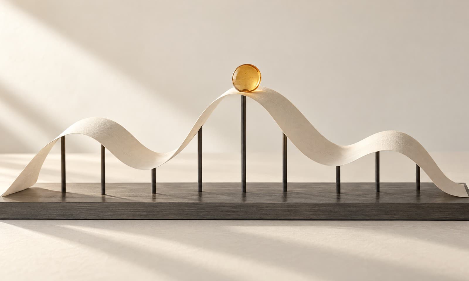 A long strip of ivory paper folded into a soft wave over a graphite plate, with thin charcoal rods beneath it and an amber disc on the highest crest