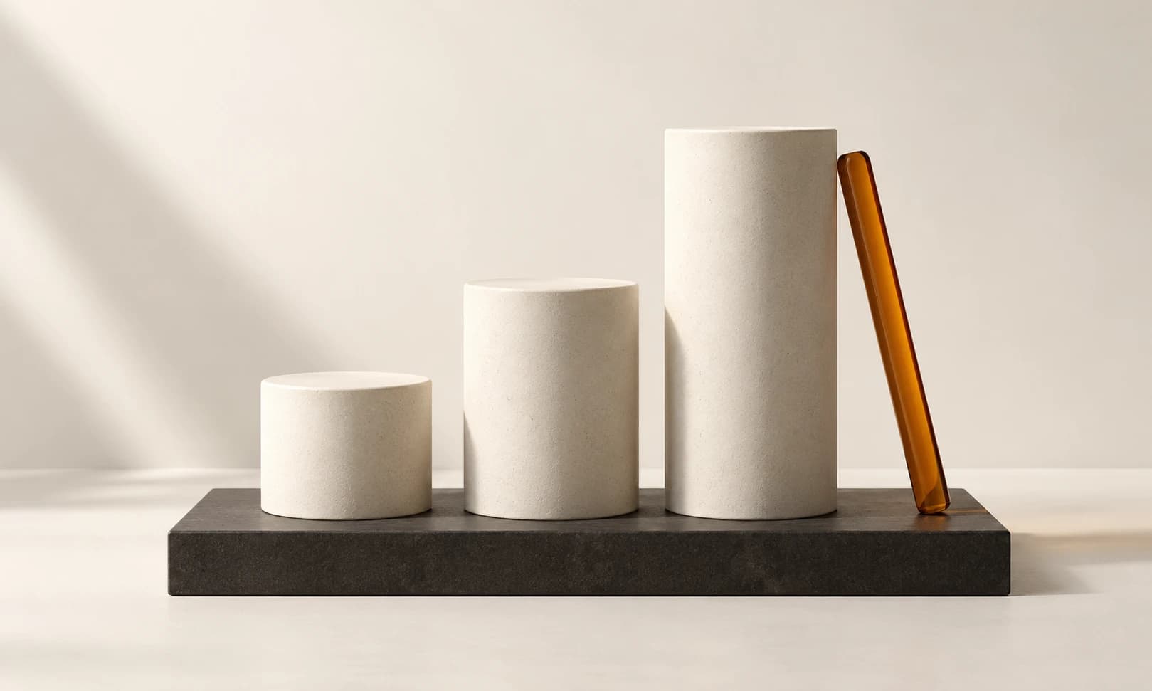 Three ascending ivory cylinders on a shared charcoal plinth beside an amber rod