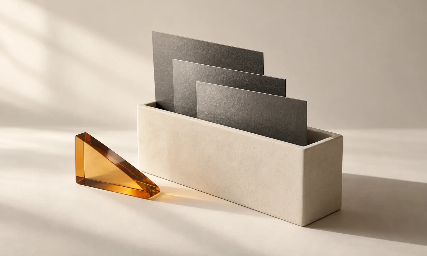 An ivory ceramic rectangular vessel with three thin graphite sheets sliding into it, one amber glass wedge beside it, suggesting delivery and careful packaging