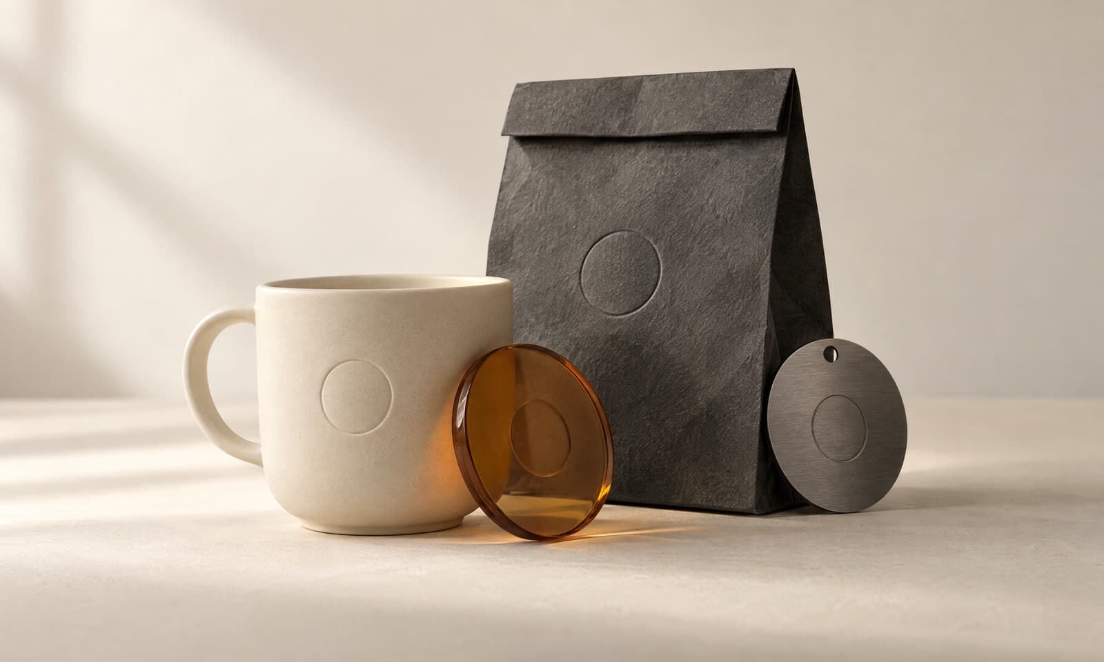 An ivory ceramic cup, a charcoal paper bag and a graphite plate, each with the same circular emboss, and an amber disc leaning on the cup